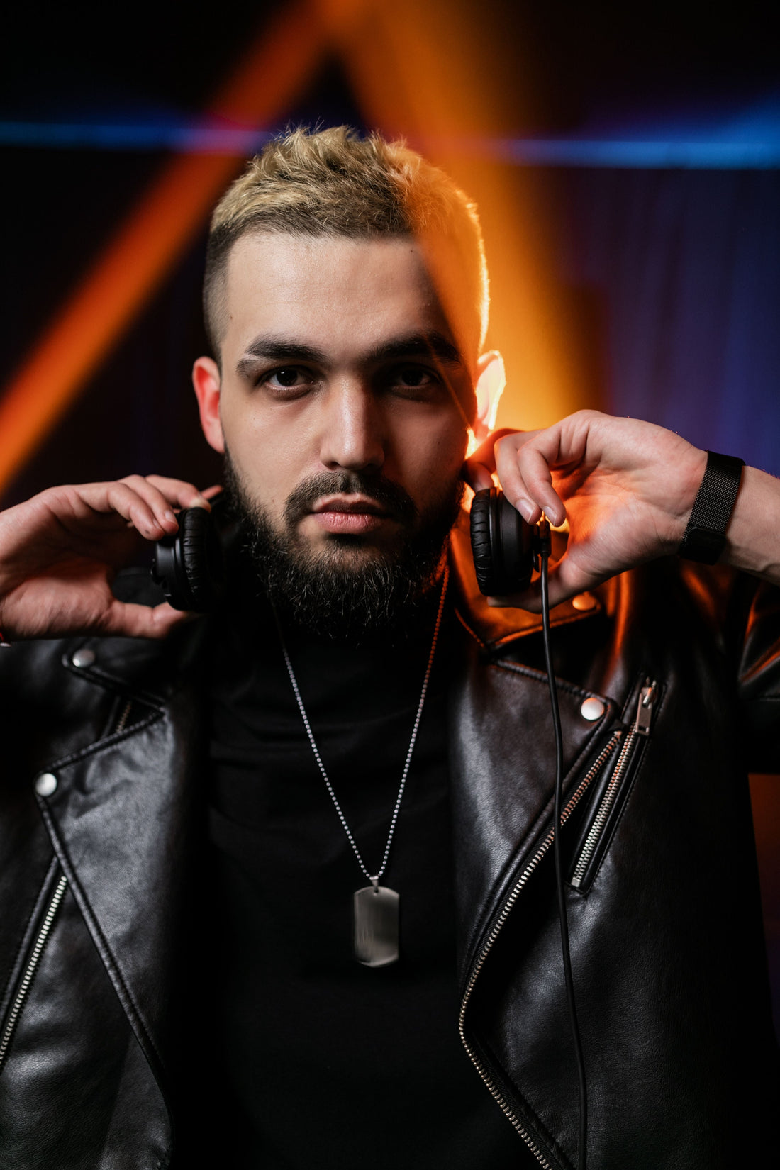 A man in a black leather jacket holding headphones around his neck under blue and orange lights.