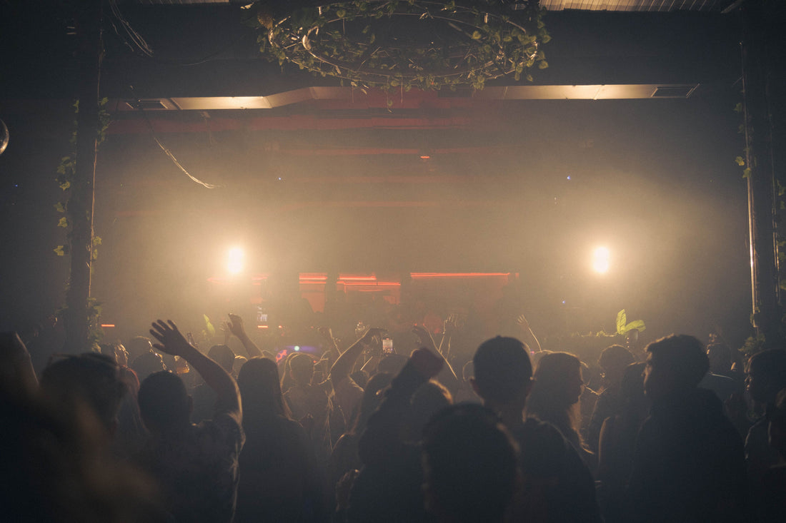 A crowd with raised hands facing bright amber stage lights in a hazy indoor venue.